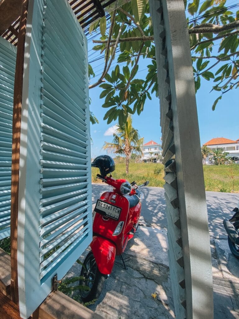Scooter parked near a traditional Balinese window in Canggu, Bali, with lush greenery and clear blue.