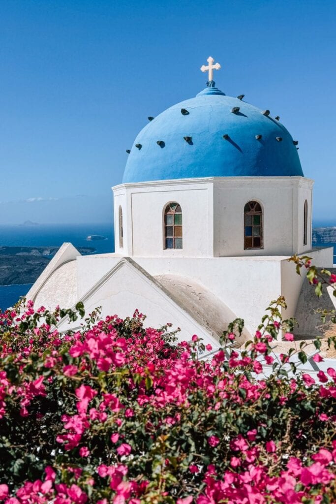 Iconic Santorini church with blue dome, surrounded by vibrant bougainvillea flowers, overlooking the Aegean Sea.