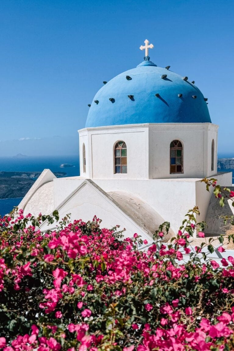 Iconic Santorini church with blue dome, surrounded by vibrant bougainvillea flowers, overlooking the Aegean Sea.