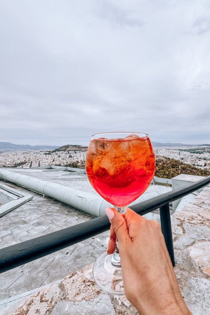 Hand holding Aperol Spritz with Athens cityscape in the background.