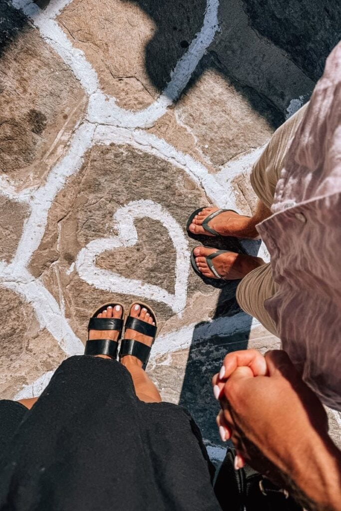 View of two people holding hands on a stone path with a heart shape drawn in white chalk, overlookin.