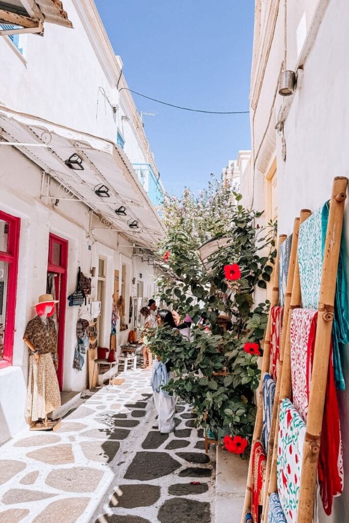 Sunlit narrow street in Paros with local shops and colorful textiles, perfect for exploring Greek is.